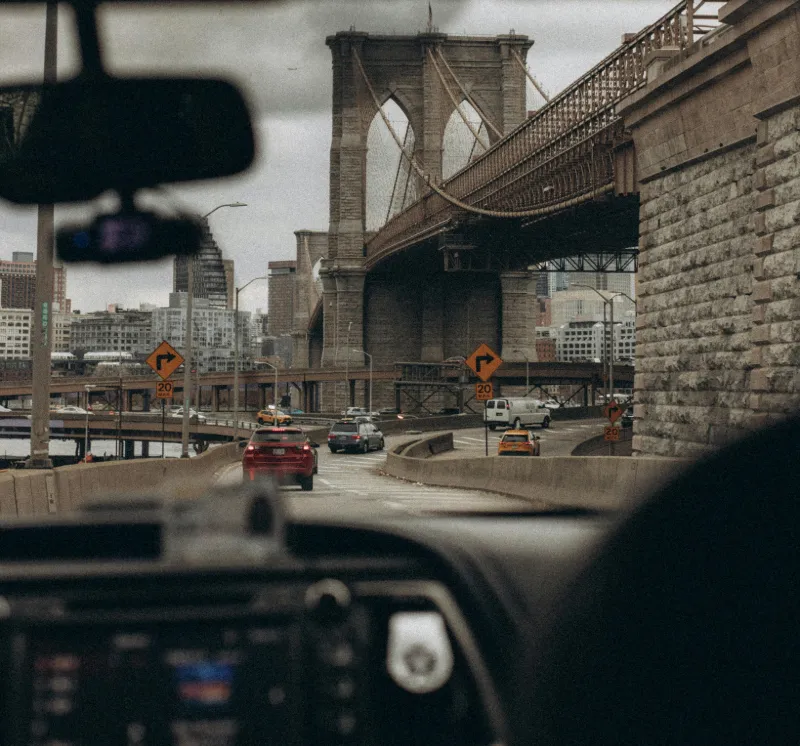 View of the Brooklyn Bridge and surrounding cityscape seen through the windshield of a car on a highway with several other cars.